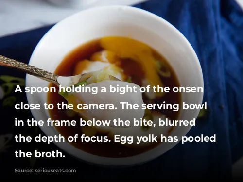 A spoon holding a bight of the onsen egg close to the camera. The serving bowl is in the frame below the bite, blurred by the depth of focus. Egg yolk has pooled into the broth.