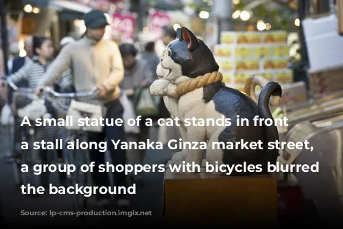 A small statue of a cat stands in front of a stall along Yanaka Ginza market street, with a group of shoppers with bicycles blurred in the background