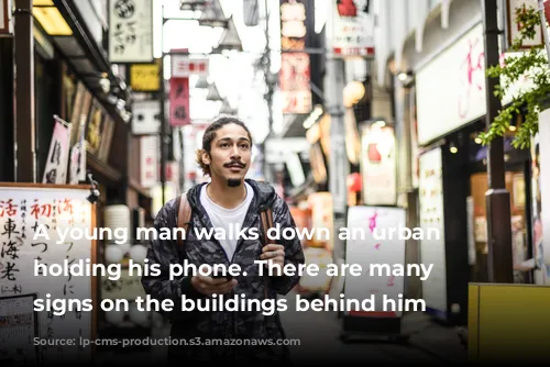 A young man walks down an urban street holding his phone. There are many Japanese signs on the buildings behind him