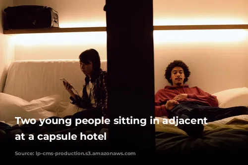 Two young people sitting in adjacent pods at a capsule hotel