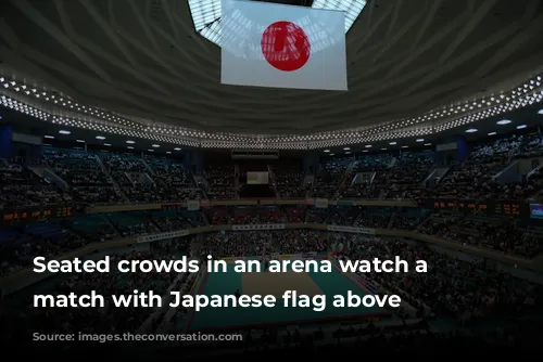 Seated crowds in an arena watch a judo match with Japanese flag above