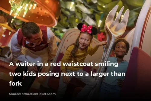 A waiter in a red waistcoat smiling at two kids posing next to a larger than life fork