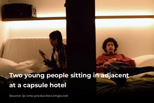 Two young people sitting in adjacent pods at a capsule hotel