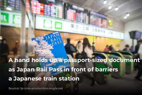 A hand holds up a passport-sized document labeled as Japan Rail Pass in front of barriers at a Japanese train station