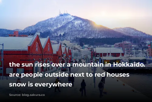 the sun rises over a mountain in Hokkaido. There are people outside next to red houses and snow is everywhere